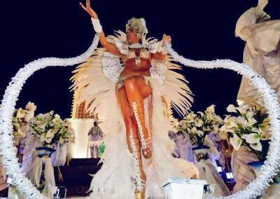 A performer in an elaborate white feathered costume with a jeweled headdress dances atop a float during a nighttime Brazilian Carnival parade.